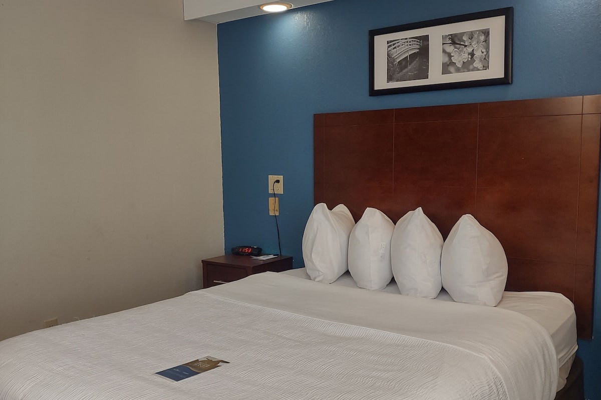 Quarto de hotel com uma cama grande, cabeceira de madeira, mesa de cabeceira com um relógio e um quadro na parede.