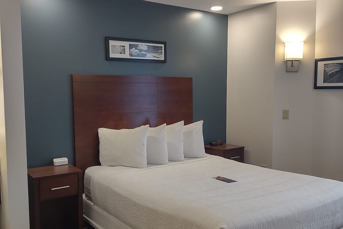 Cama bem arrumada com lençóis brancos em quarto com parede azul, cabeceira de madeira e duas mesas de cabeceira com abajures.
