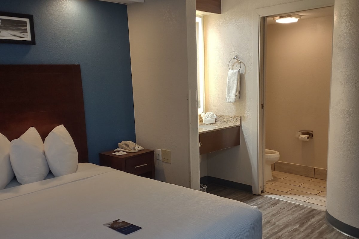 Quarto de hotel com uma cama grande, mesa de cabeceira, banheiro visível através de uma porta aberta e cabeceira de madeira.