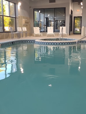 Piscina interna com água cristalina, rodeada por espreguiçadeiras e uma parede de vidro.