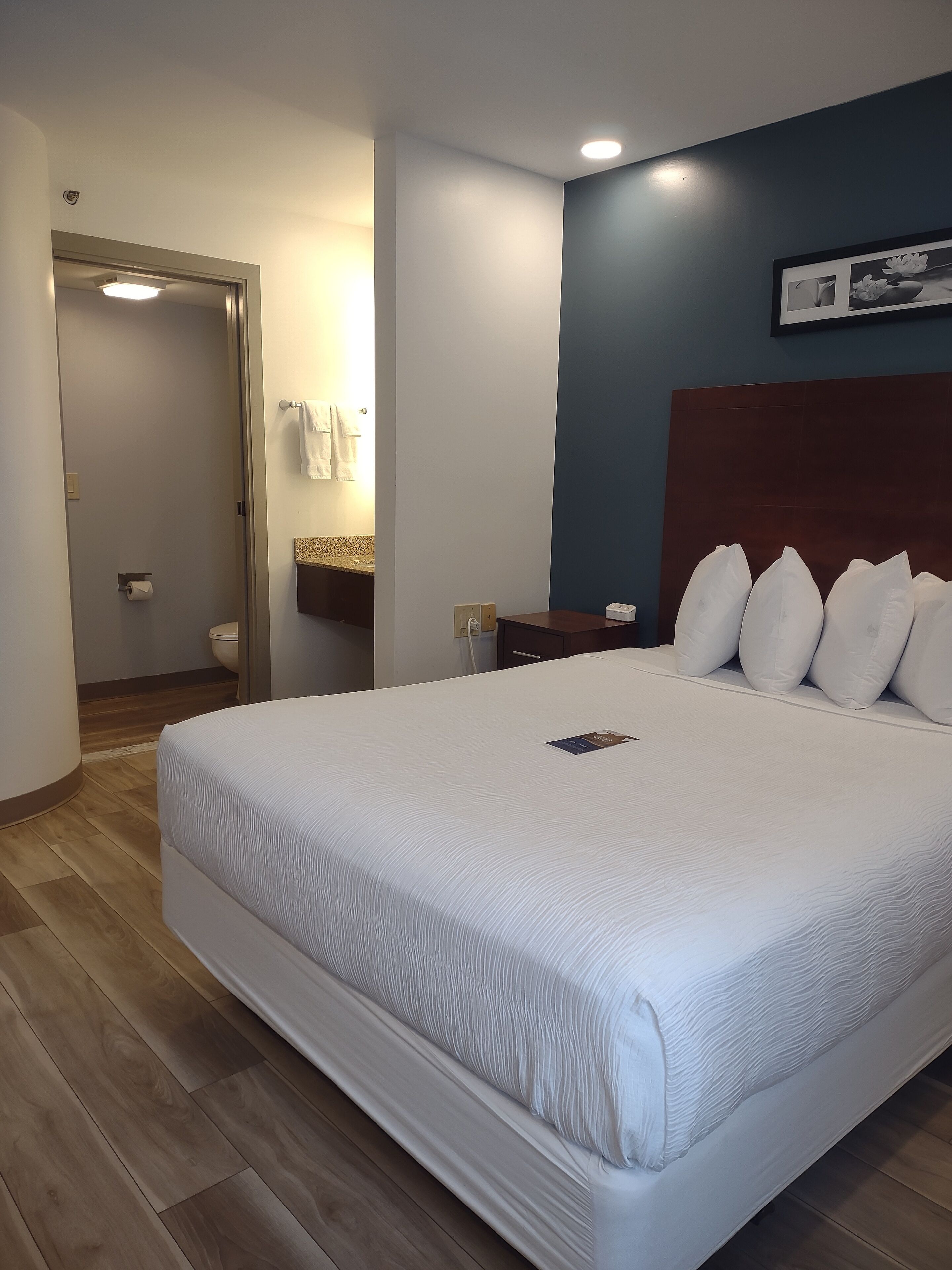 A hotel room with a large bed, a wooden floor, a bathroom with a towel rack, and a nightstand with a phone.
