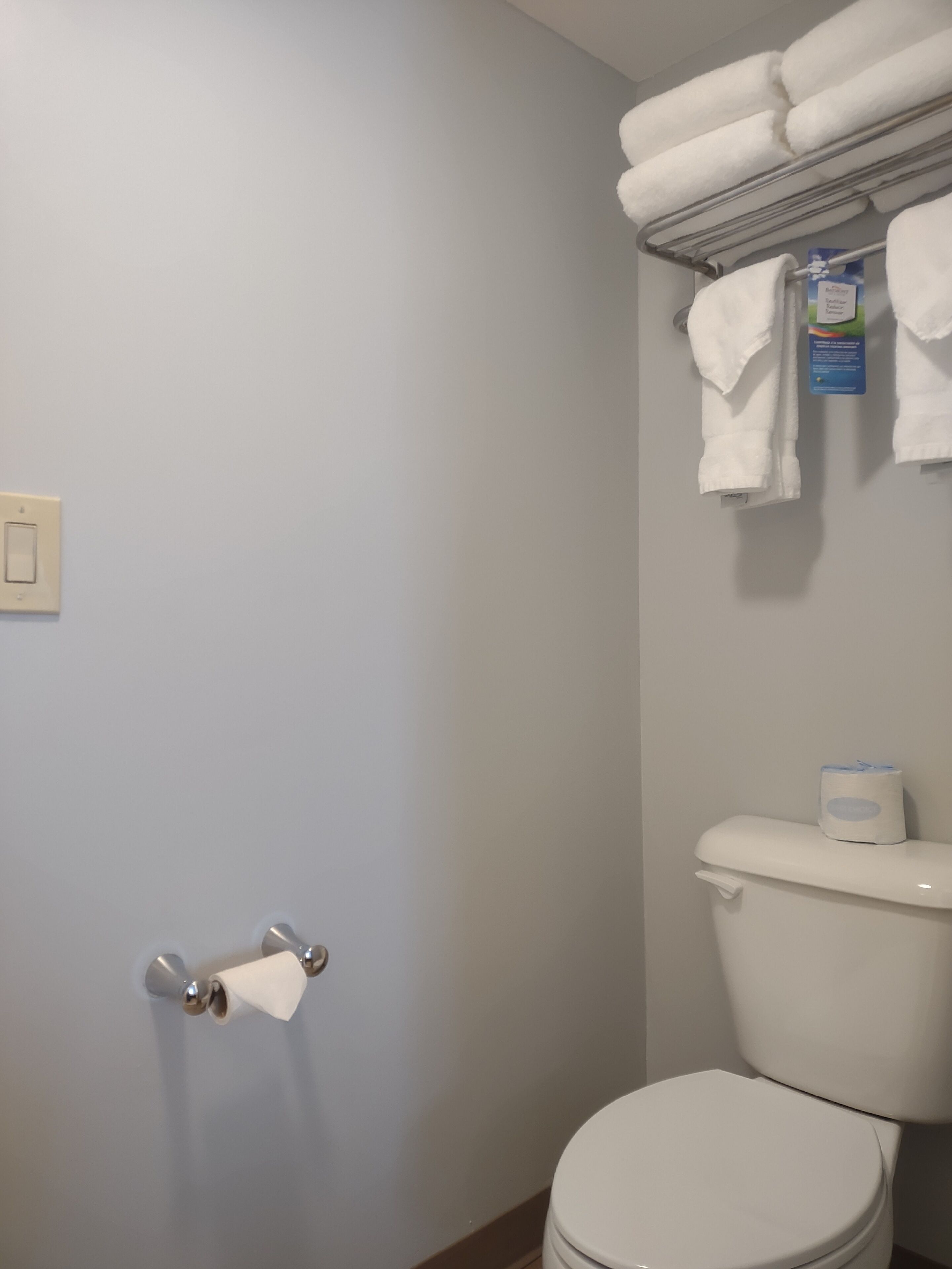 A bathroom with a toilet, a towel rack with folded towels, a toilet paper holder, and a light switch on the wall.