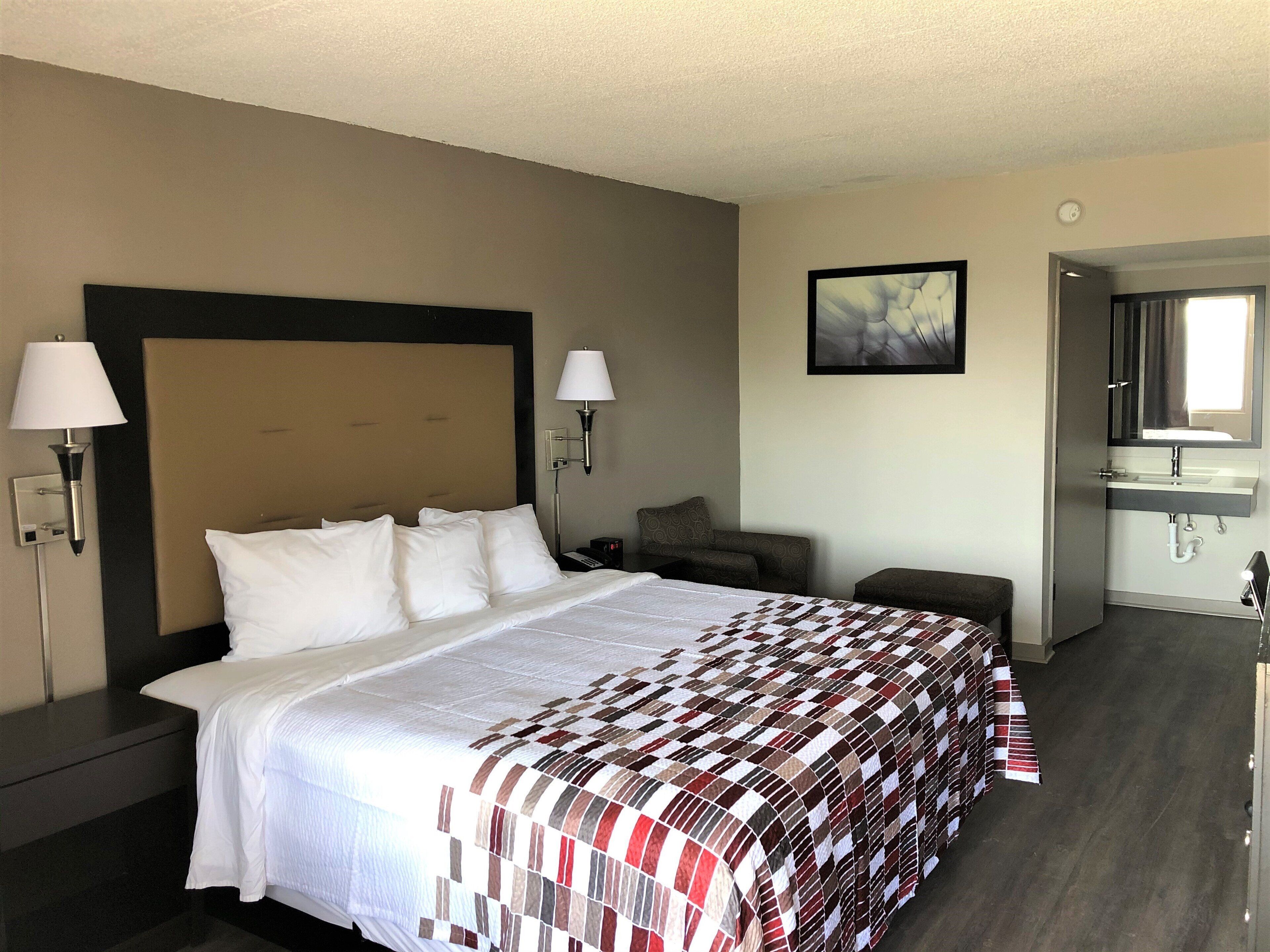 Une chambre d’hôtel avec un grand lit, deux lampes de chevet, un petit coin salon, un tableau au mur et une salle de bain avec un lavabo et un miroir.