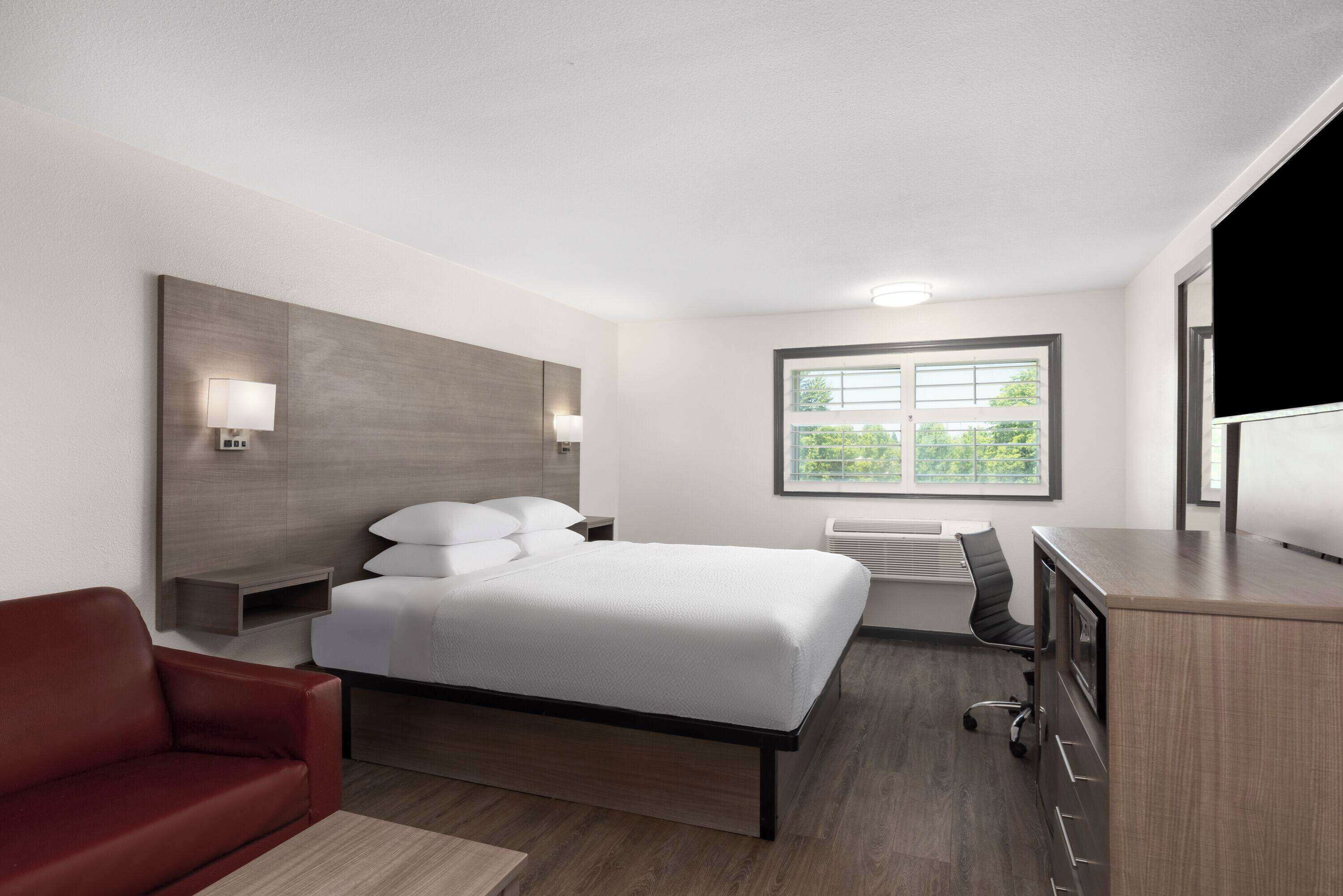 A modern hotel room with a large bed, a desk, a chair, a red sofa, and a window with a view of greenery.