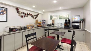 A clean, modern kitchen with a central island, equipped with a coffee machine, a toaster, and various food items. There are two dining tables with chairs, and a refrigerator is visible in the background.