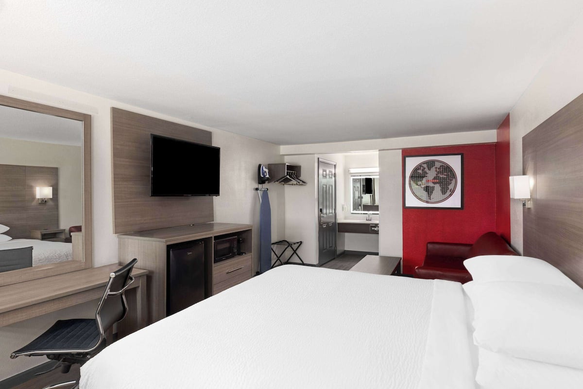 Ein Hotelzimmer mit Bett, Schreibtisch, Fernseher, Spiegel, einer roten Akzentwand und einer Tür zu einem Badezimmer.