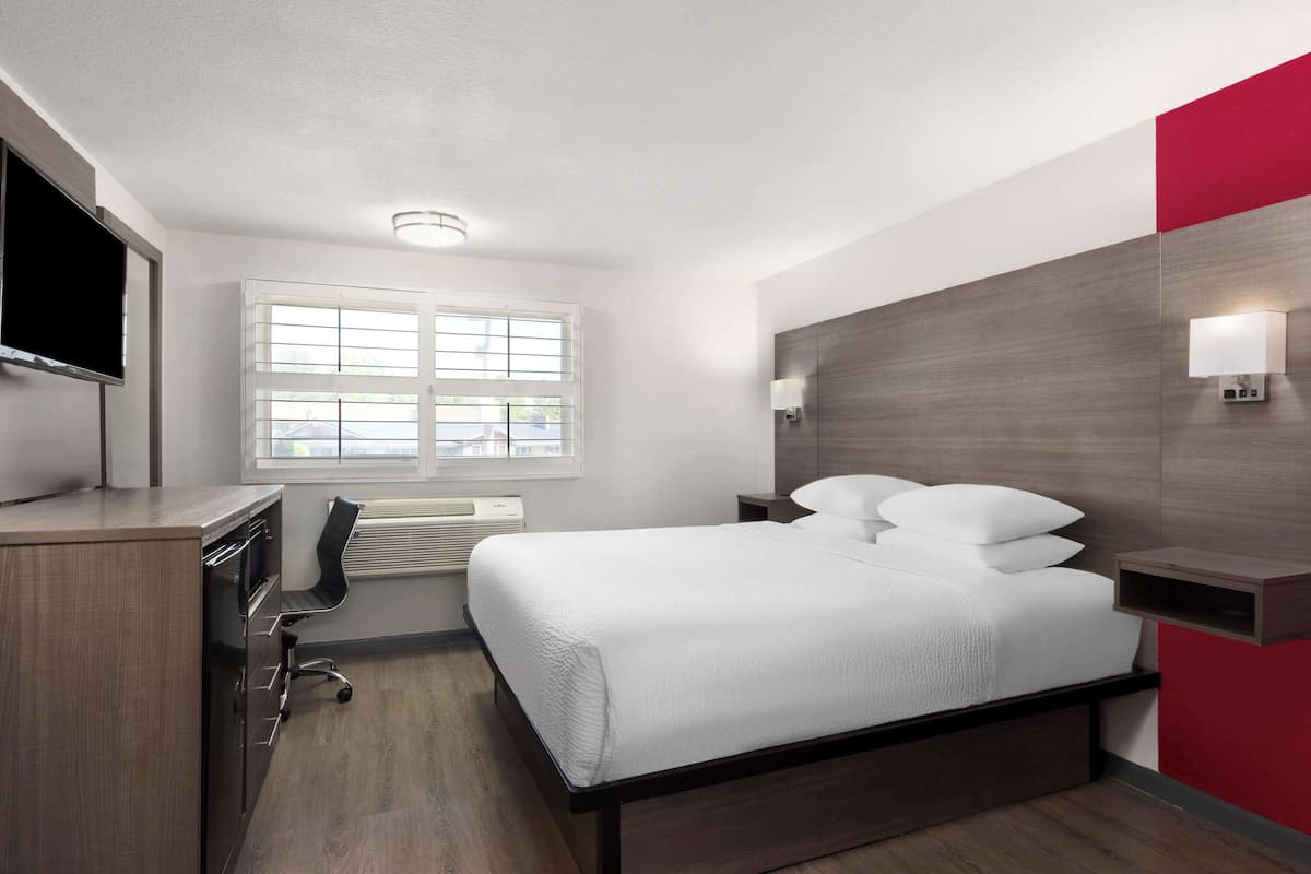 Ein modernes Hotelzimmer mit Bett, Schreibtisch, Stuhl, Fernseher, Fenster und Wandlampe.