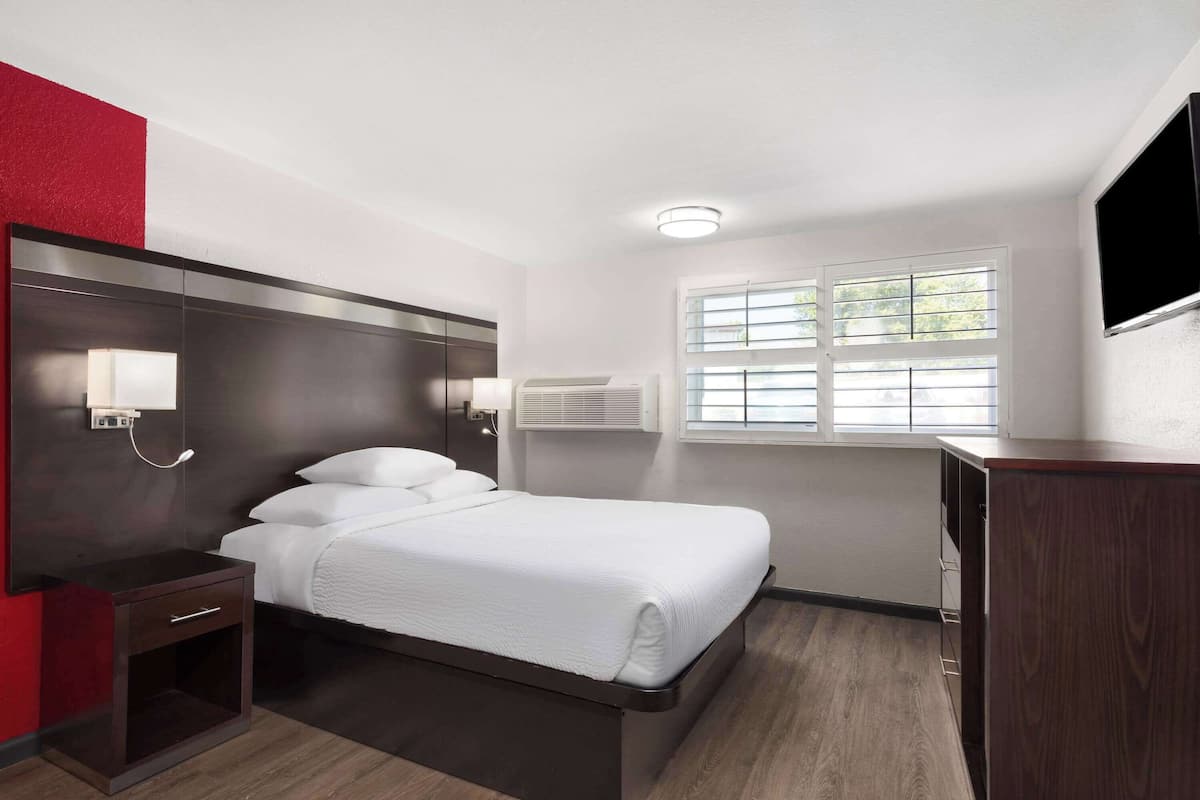 Ein Hotelzimmer mit Bett, Nachttisch, Lampe, Klimaanlage, Fenster und einem Fernseher an der Wand.
