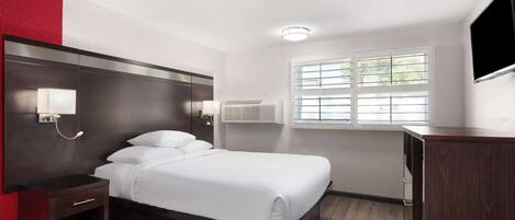 A hotel room with a bed, bedside table, lamp, air conditioning unit, window, and a TV on the wall.
