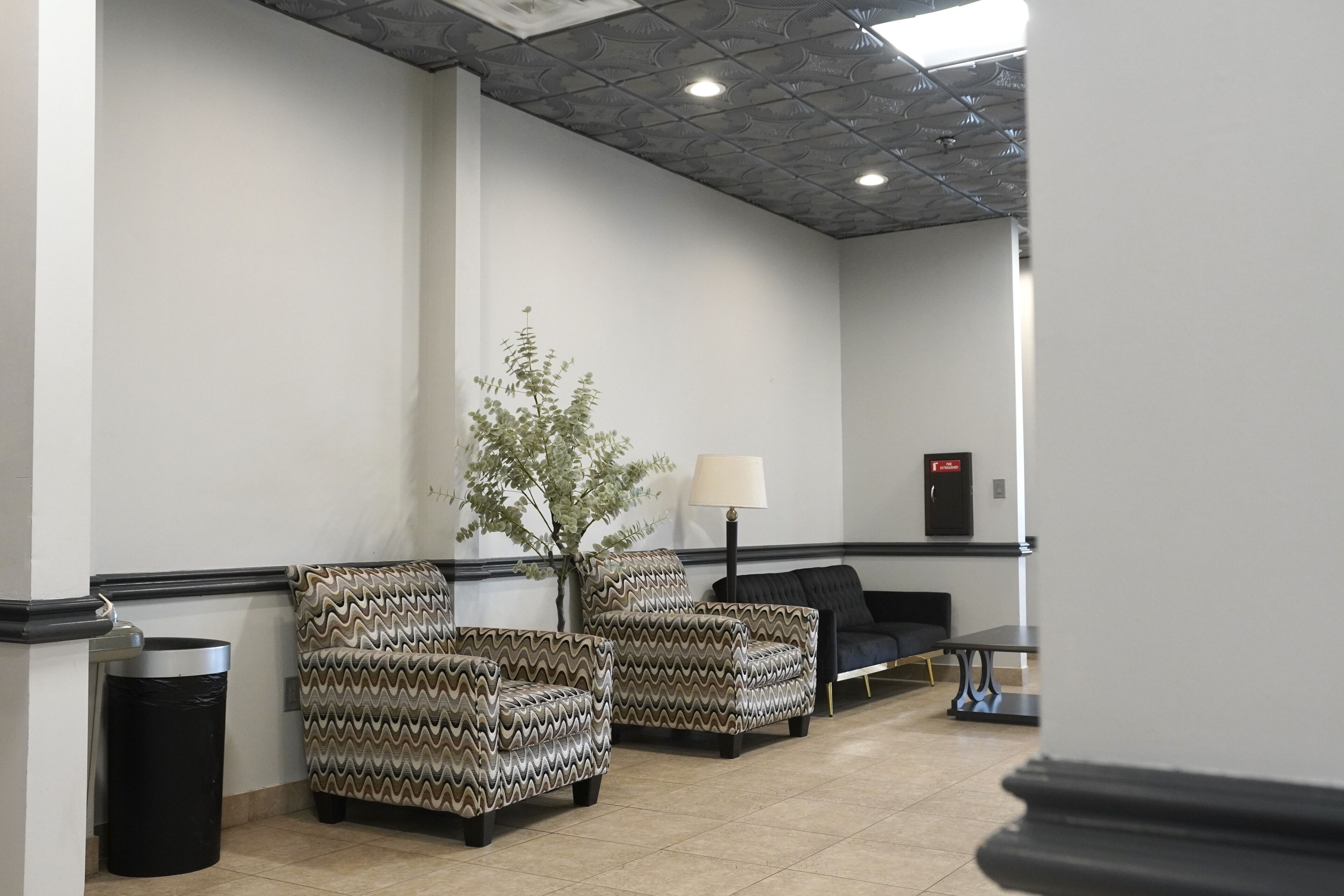 A waiting area with two patterned armchairs, a small table, a lamp, and a plant.