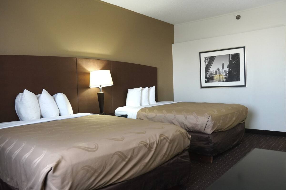 A hotel room with two beds, a nightstand with a lamp, a framed cityscape picture on the wall, and a dark floor.