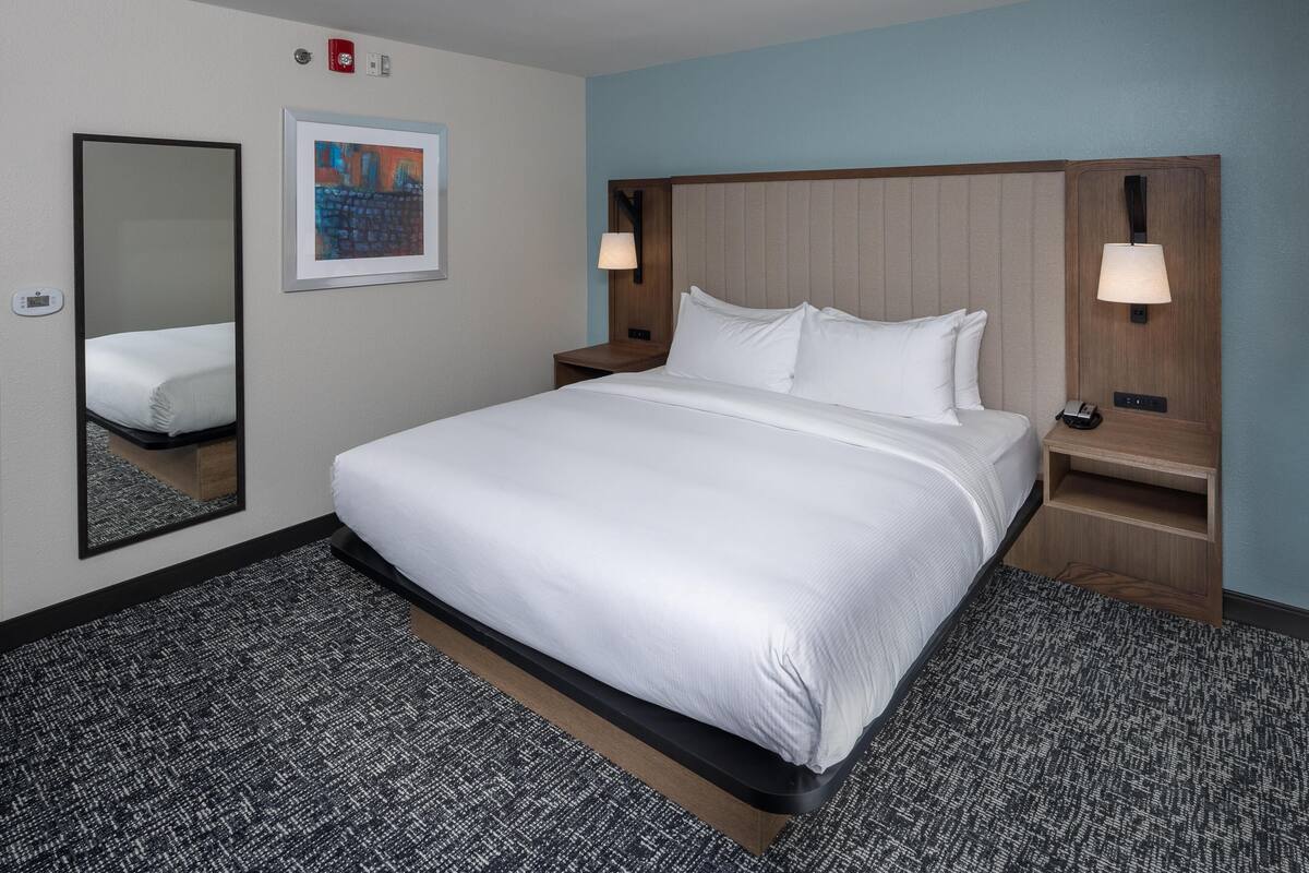 A hotel room with a large bed, a mirror, a framed picture, two bedside tables with lamps, and a carpeted floor.