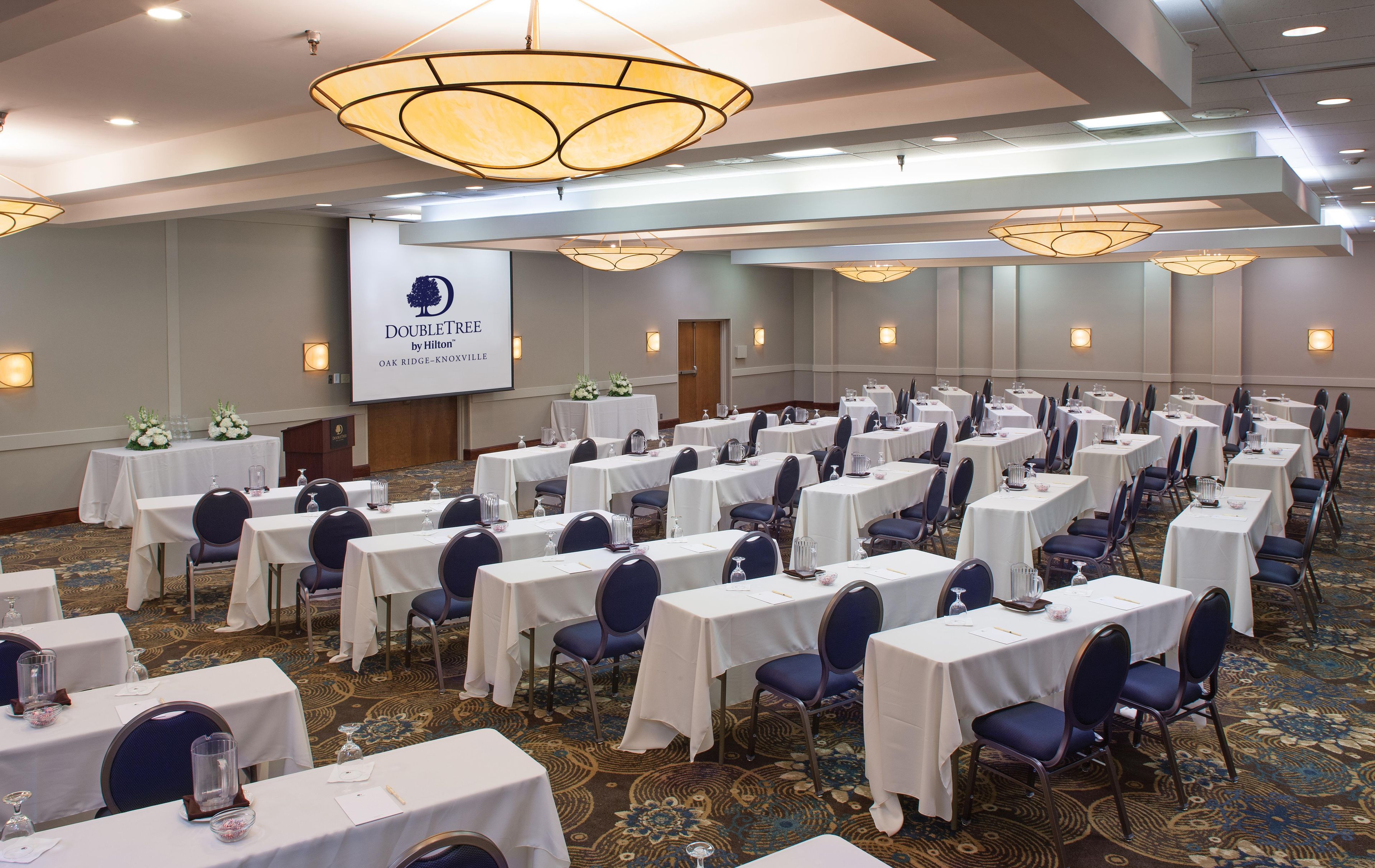 A conference room with multiple tables, chairs, and a large screen displaying the DoubleTree by Hilton logo.