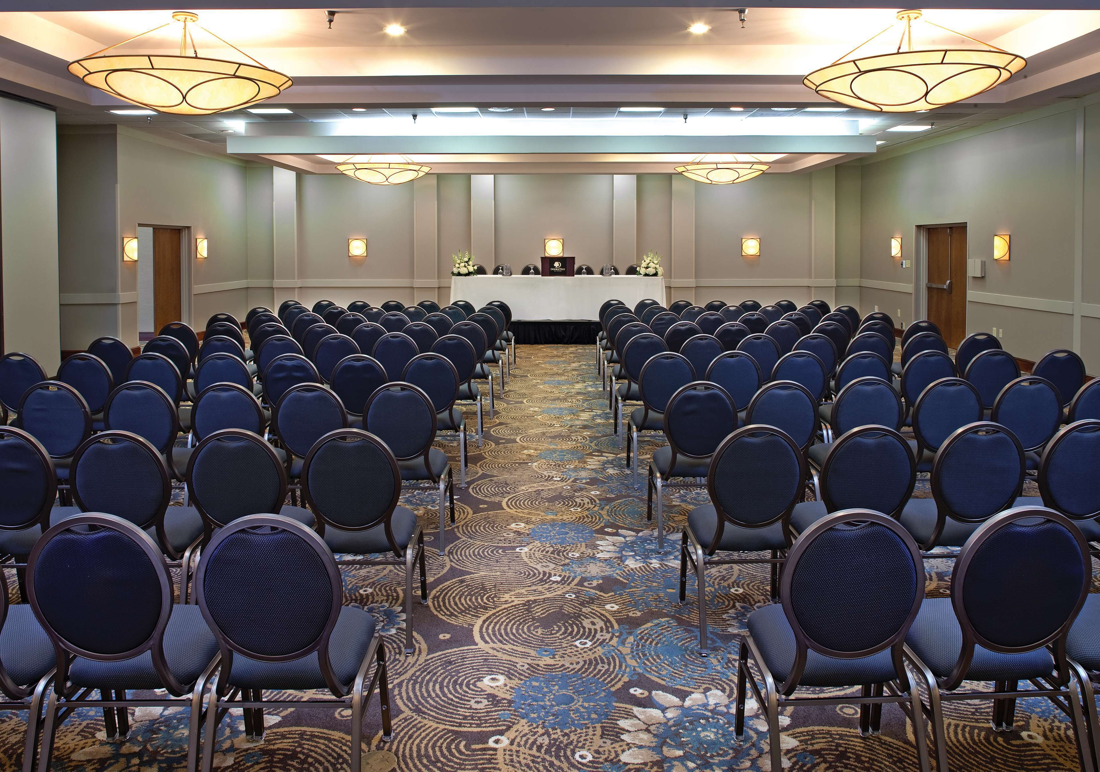 A conference room with rows of chairs facing a table with a podium, equipped with a carpeted floor and wall-mounted lighting.