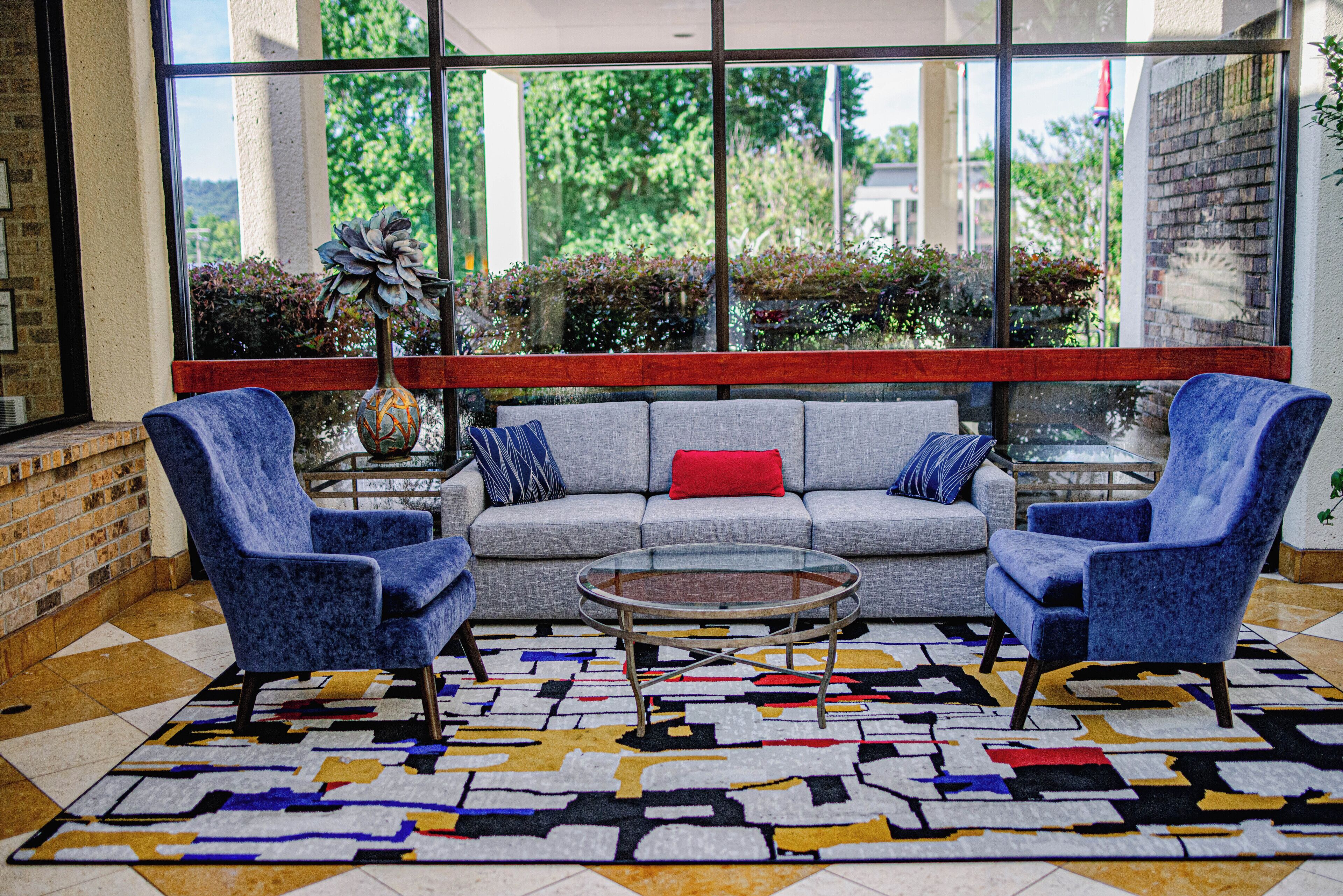 Een moderne zithoek met een grijze bank, twee blauwe fauteuils, een glazen salontafel en een vloerkleed met een patroon.