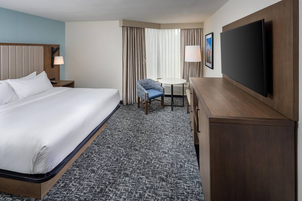 A hotel room with a bed, a desk, a chair, a TV, and a lamp.
