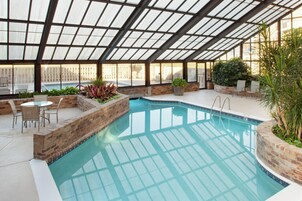 Una piscina interior con techo de cristal, rodeada de paredes de ladrillo y amueblada con sillas y plantas en macetas.