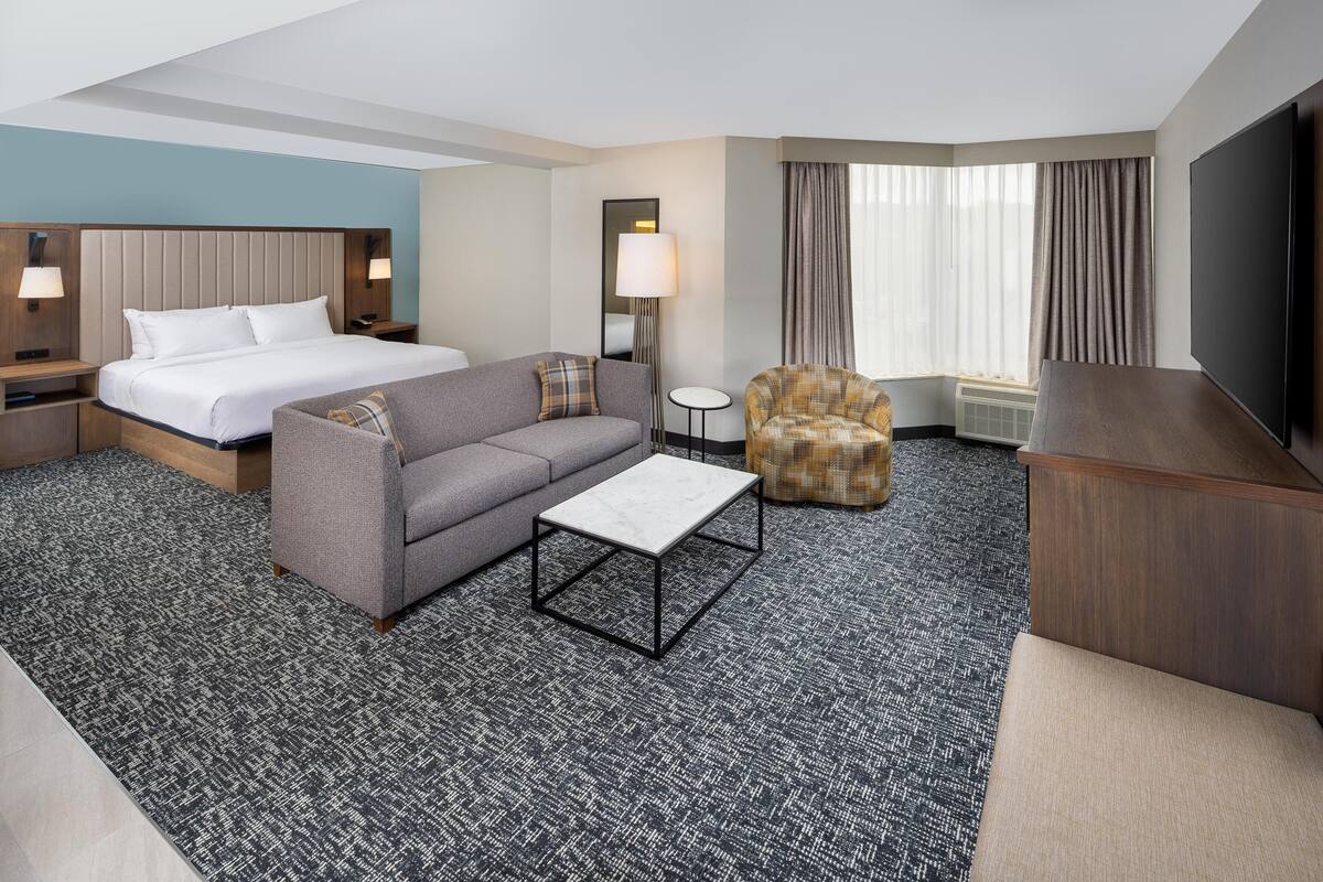 A hotel room with a bed, a sofa, a chair, a coffee table, a TV, and a lamp.