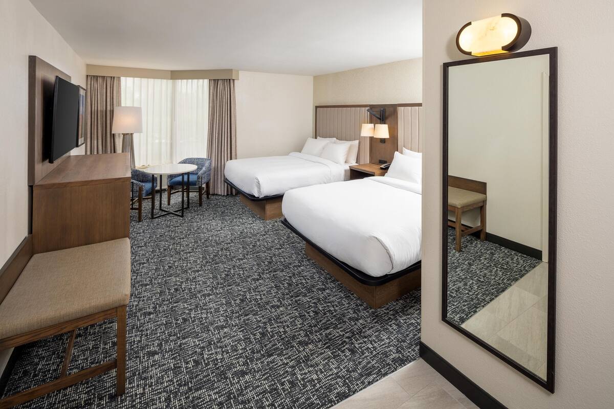 A hotel room with two beds, a desk, a chair, a TV, and a large mirror.