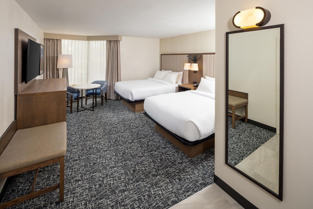 A hotel room with two beds, a desk, a chair, a TV, and a large mirror.