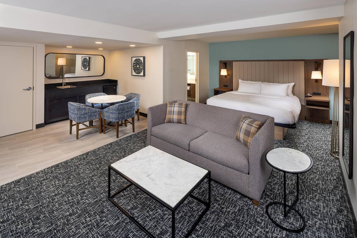 A hotel room with a bed, a sofa, a round table, a small round table, a desk, a chair, a mirror, and a wall-mounted lamp.
