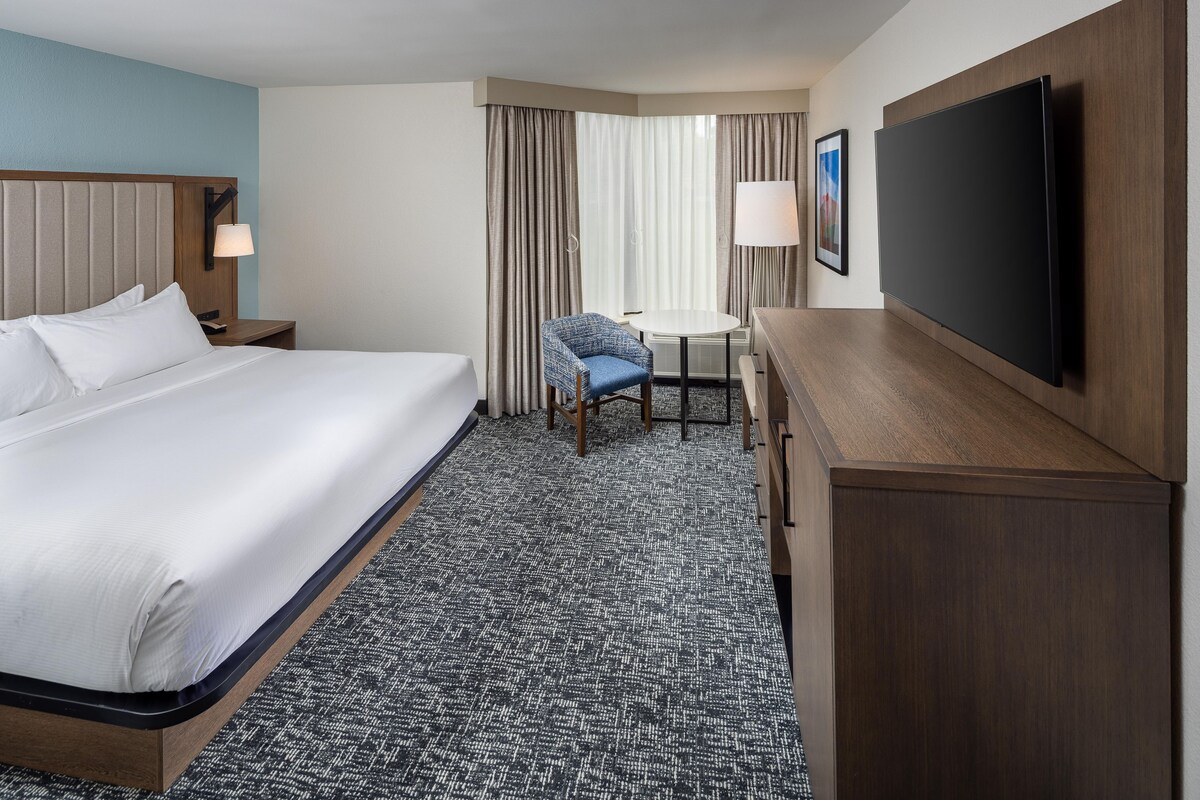 A hotel room with a bed, a desk, a chair, a TV, and a lamp.