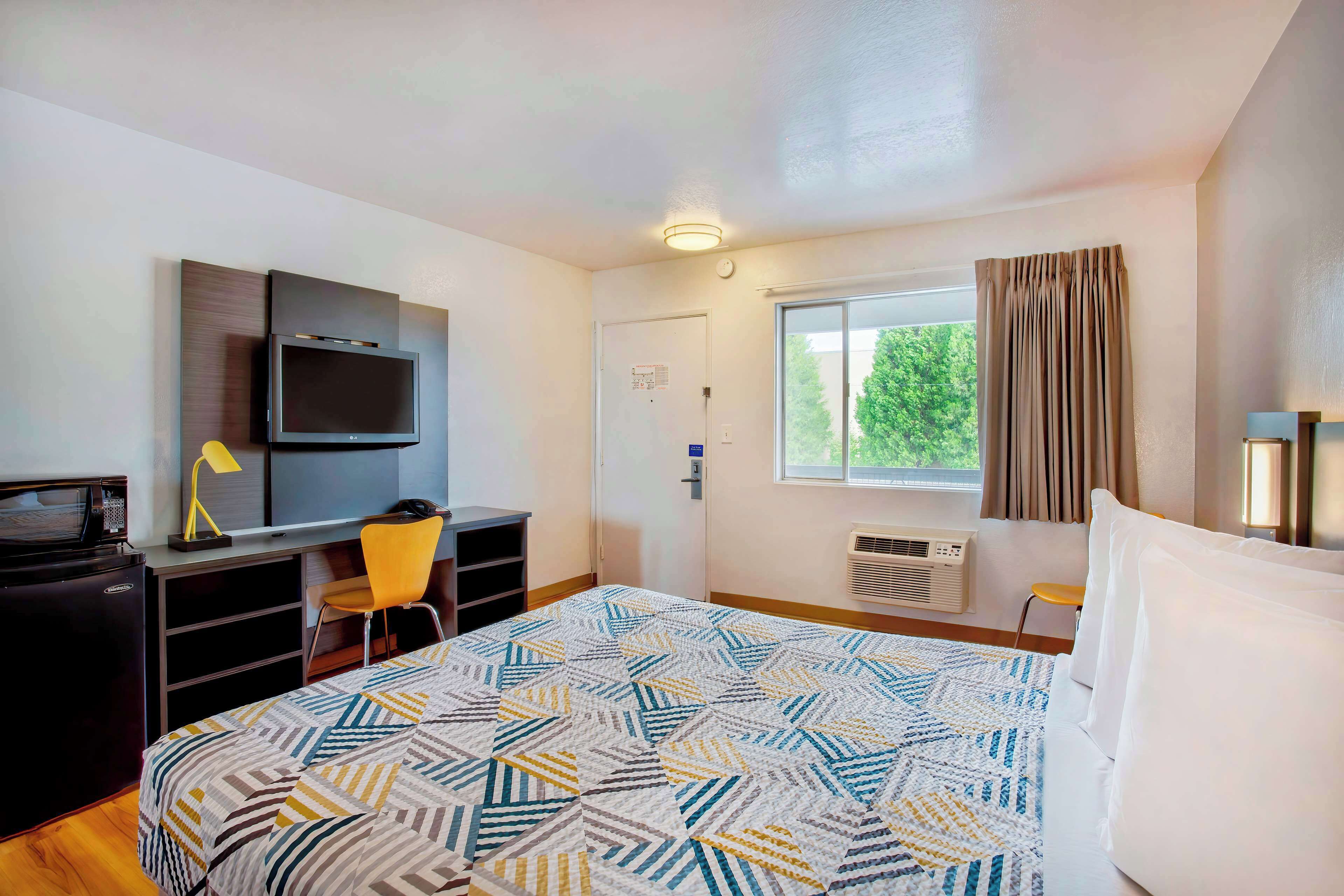 A hotel room with a bed, a desk with a computer, a TV, and a window with curtains.