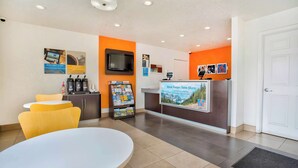 Un espace d’accueil d’hôtel avec un comptoir de réception blanc, un téléviseur, une machine à café et un présentoir à magazines.