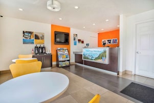 A hotel reception area with a white reception desk, a TV, a coffee machine, and a magazine rack.