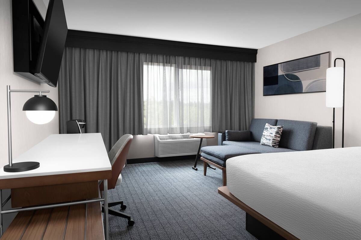 A hotel room with a bed, a desk, a chair, a sofa, a TV, and a window with curtains.