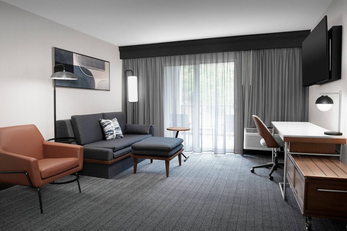 A modern hotel room with a sofa, armchair, desk, and a flat-screen TV.