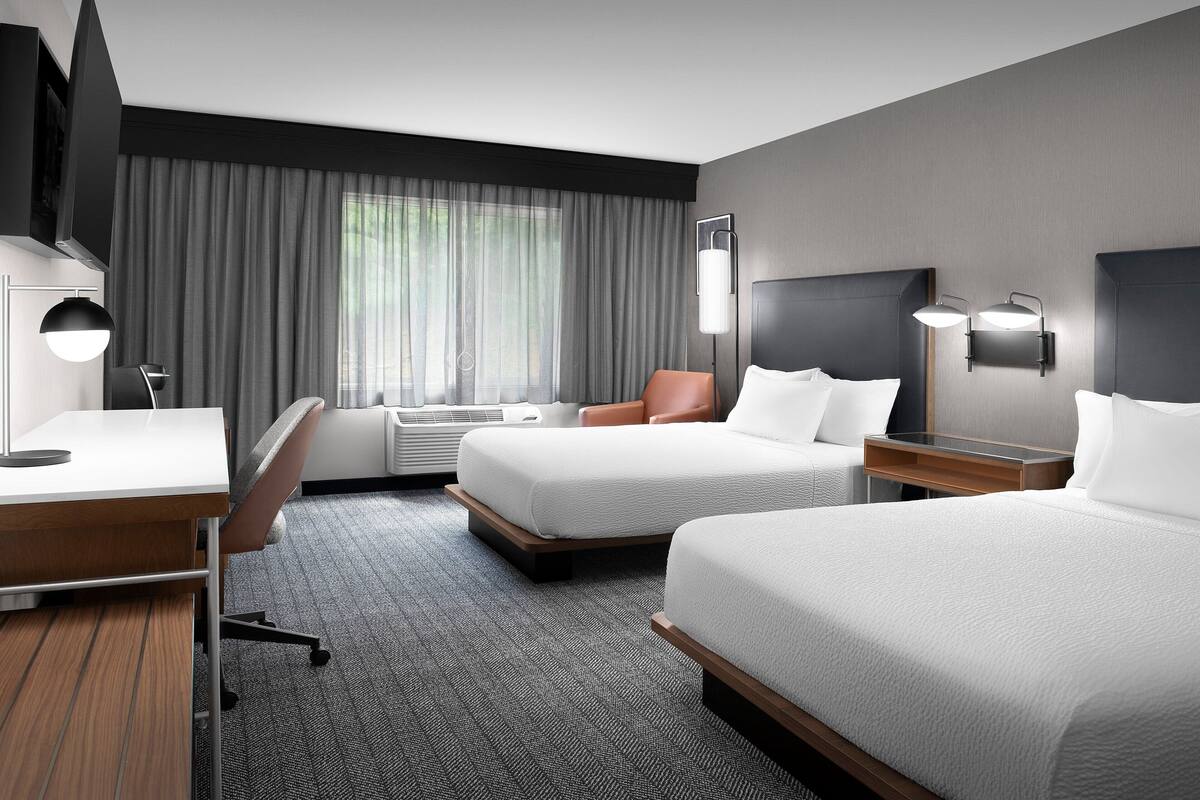 A hotel room with two beds, a desk, a chair, a TV, and a large window with curtains.