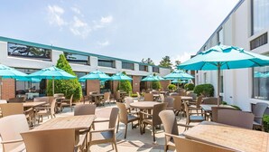An outdoor patio area with wicker tables and chairs, blue umbrellas, and a modern building in the background. - Country Inn & Suites by Radisson, Traverse City, MI (Traverse City)