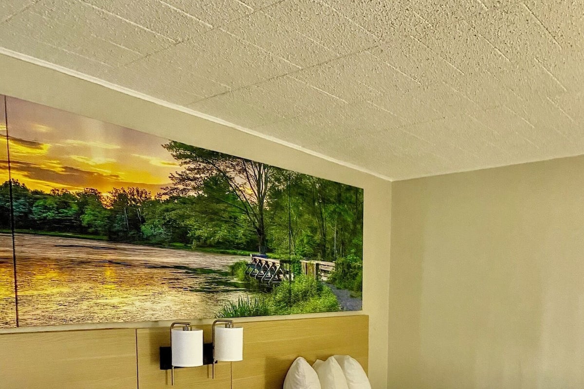 A hotel room with two beds, a painting of a sunset landscape, a bedside table with a lamp, and a phone.