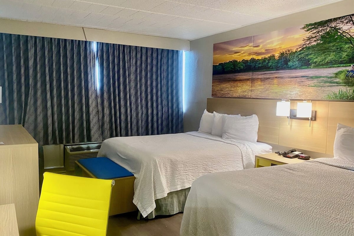 A hotel room with two beds, a desk, a chair, and a painting of a sunset over a lake.
