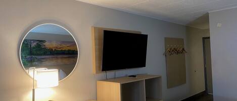 A hotel room with a TV, a mirror, a desk with a lamp, a chair, and a view of a sunset through the window.