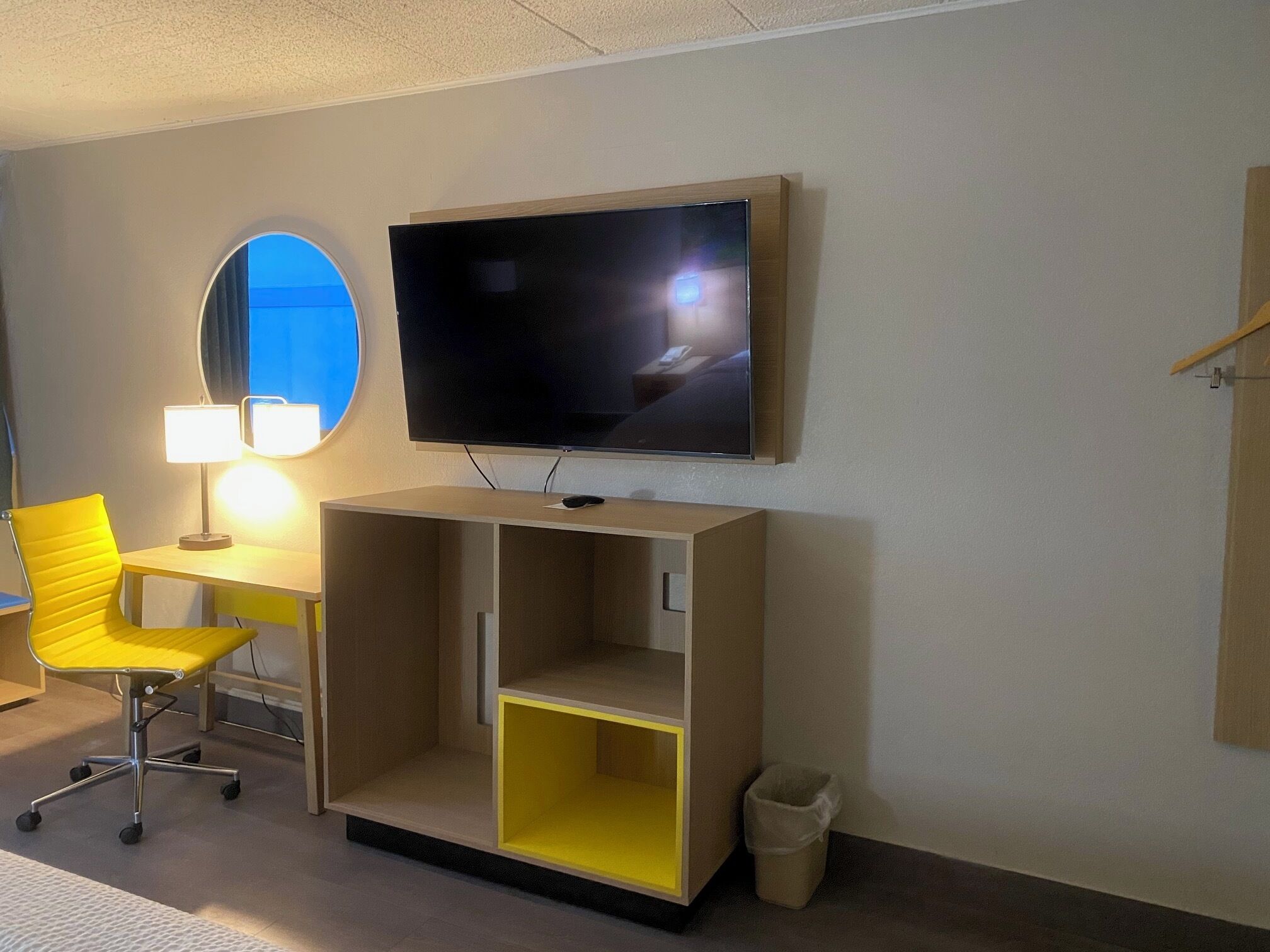 Habitación de hotel con una televisión montada en la pared, una silla amarilla, un escritorio con una lámpara y un cubo de basura pequeño.
