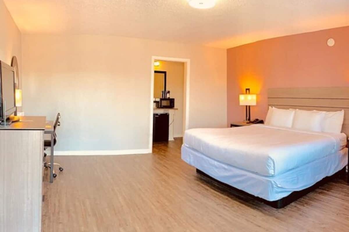 Une chambre d’hôtel avec un grand lit, un bureau avec une chaise, une table de chevet avec une lampe, et une salle de bain visible à travers une porte ouverte.