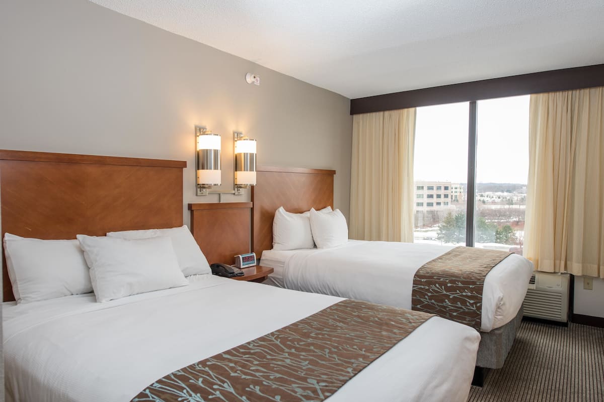 A hotel room with two beds, a wooden headboard, a nightstand with a phone, and a window with curtains.