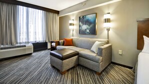 Une chambre d’hôtel avec un canapé d’angle, une petite table, une lampe fixée au mur et une grande fenêtre avec des rideaux.