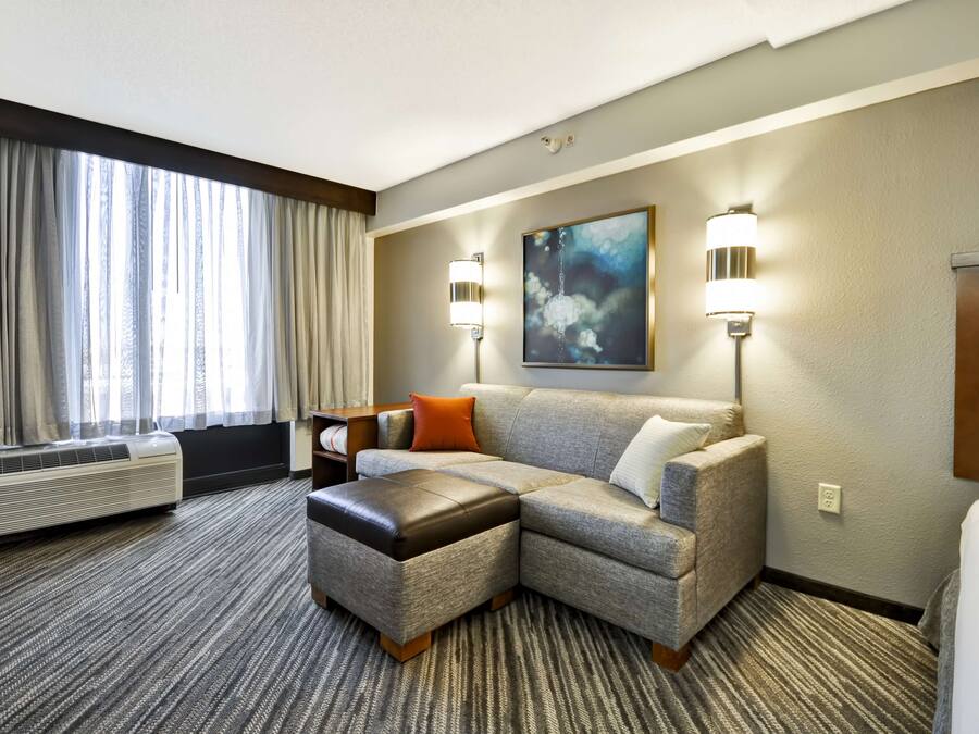 Habitación de hotel con sofá seccional, una mesita, lámpara de pared y un gran ventanal con cortinas.