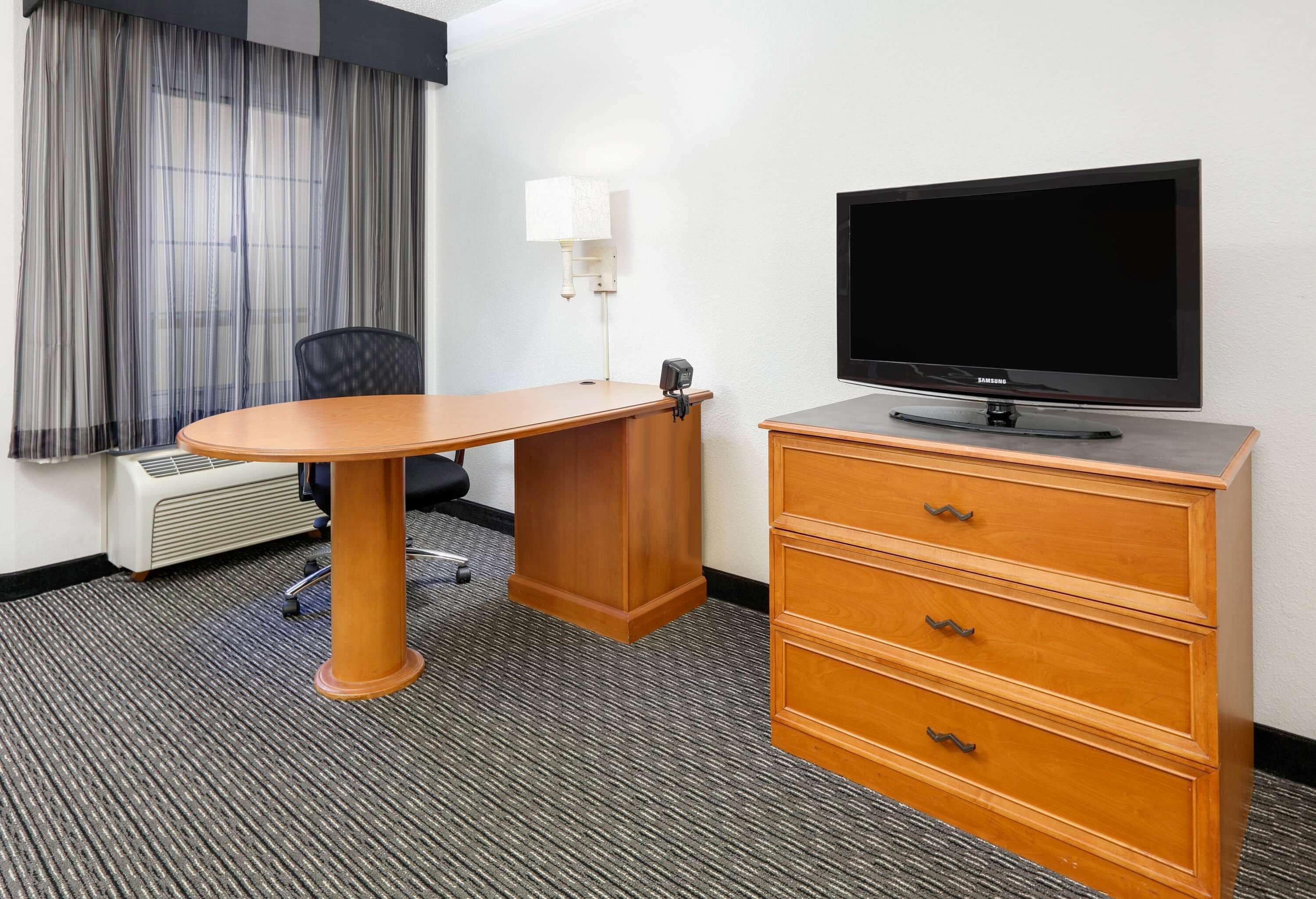 Ein Hotelzimmer mit Schreibtisch aus Holz, Flachbildfernseher, Lampe, Stuhl und Kommode.