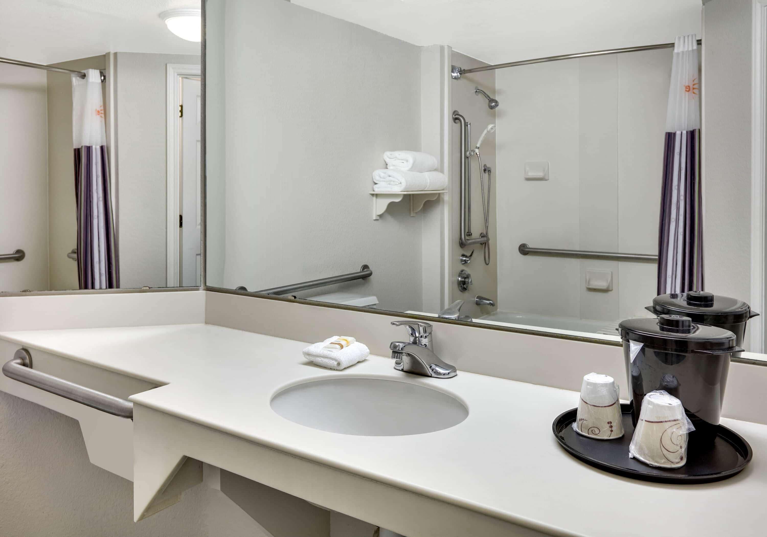 Un bagno d'hotel con lavandino, specchio, porta asciugamani, doccia e macchina del caffè sul ripiano.