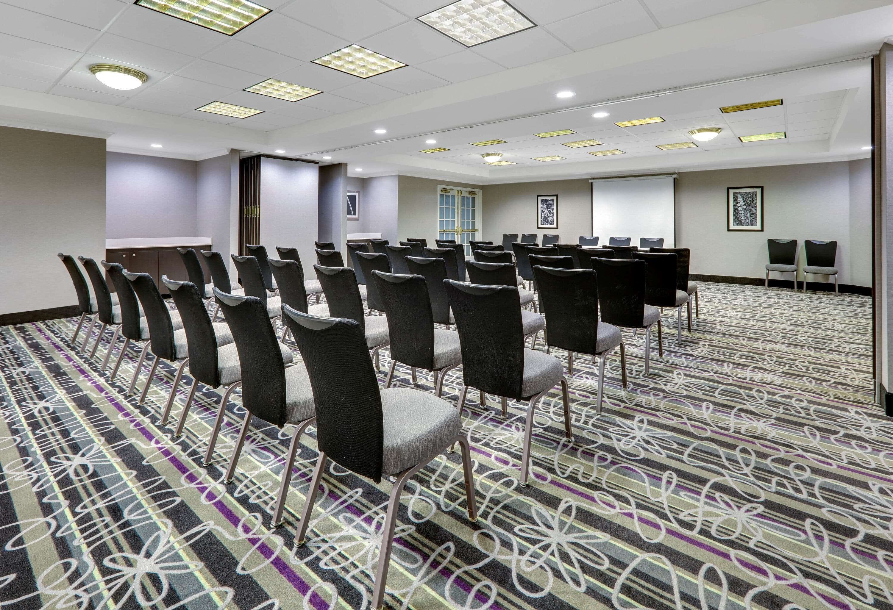 Un salón de conferencias con filas de sillas, piso alfombrado y una pantalla para proyector.