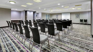 Un salón de conferencias con filas de sillas, piso alfombrado y una pantalla para proyector.