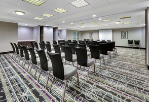 Uma sala de conferências com fileiras de cadeiras, piso coberto por carpete e uma tela de projetor.