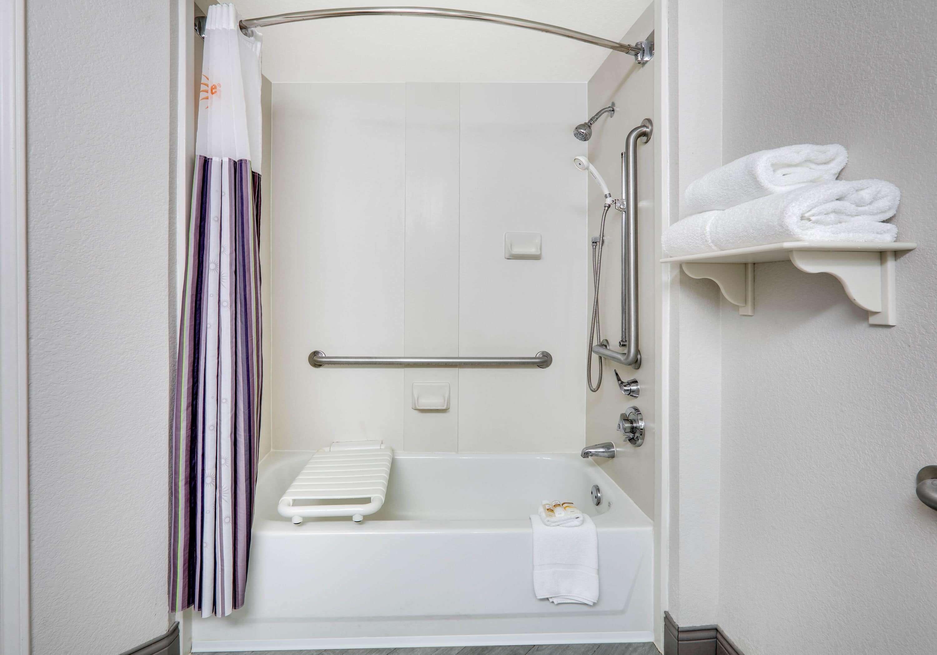 Une salle de bain avec une douche à l’italienne, des barres d ’appui, une baignoire blanche et un porte-serviettes avec des serviettes pliées.
