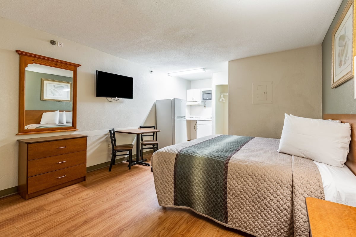 A hotel room with a bed, a desk, a chair, a dresser, a flat-screen TV, and a kitchenette.