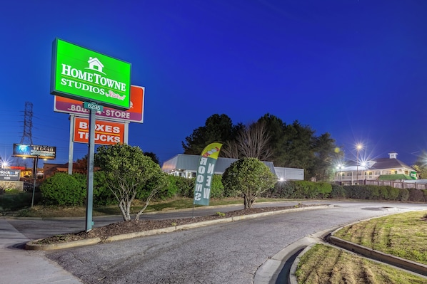 Zona comercial con un letrero verde para HomeTown Studios, un anuncio de tienda para cajas y camiones, y un letrero de servicio de seguridad.