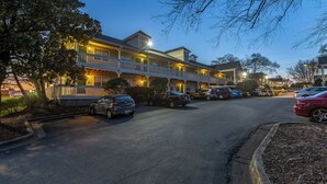 Un hotel de dos pisos con un balcón en el segundo nivel, un estacionamiento con varios coches y un cielo despejado.